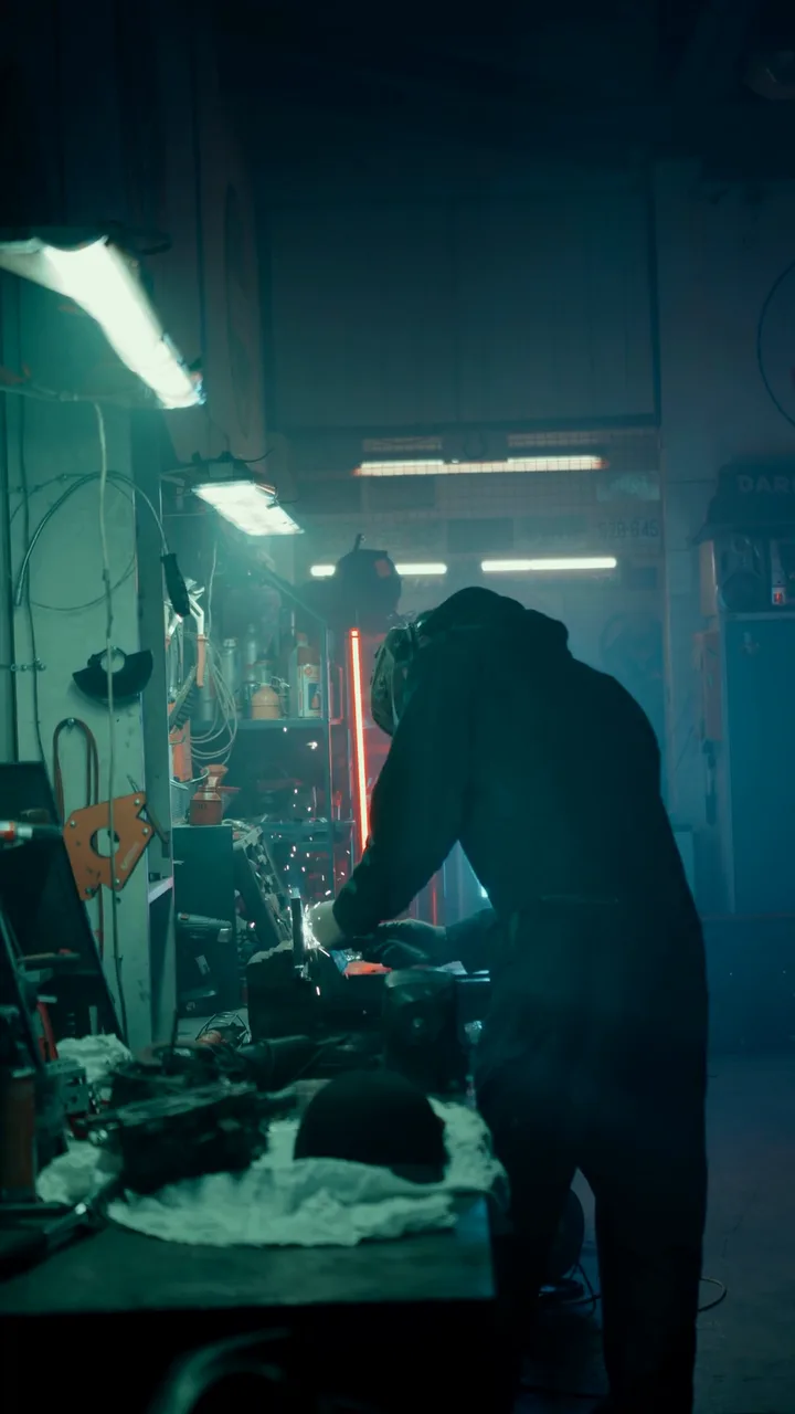 A welder bends over his workbench in a dim workshop, sparks and blue light around him.