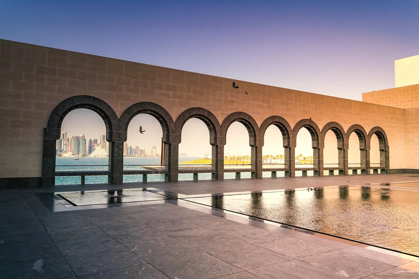 A colonnade of arches framing the Doha skyline over still water.