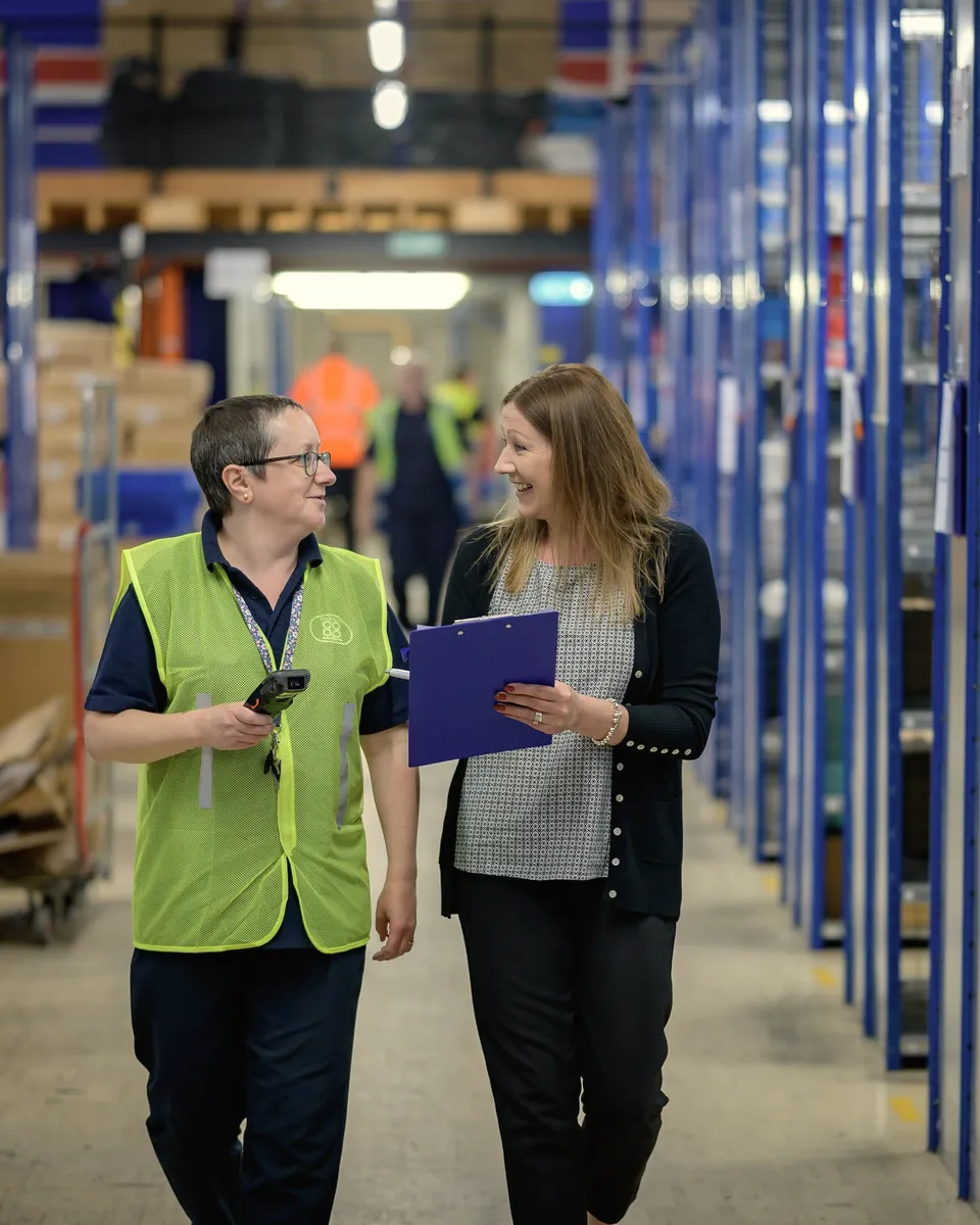 Two colleagues walk a warehouse aisle, reviewing notes.